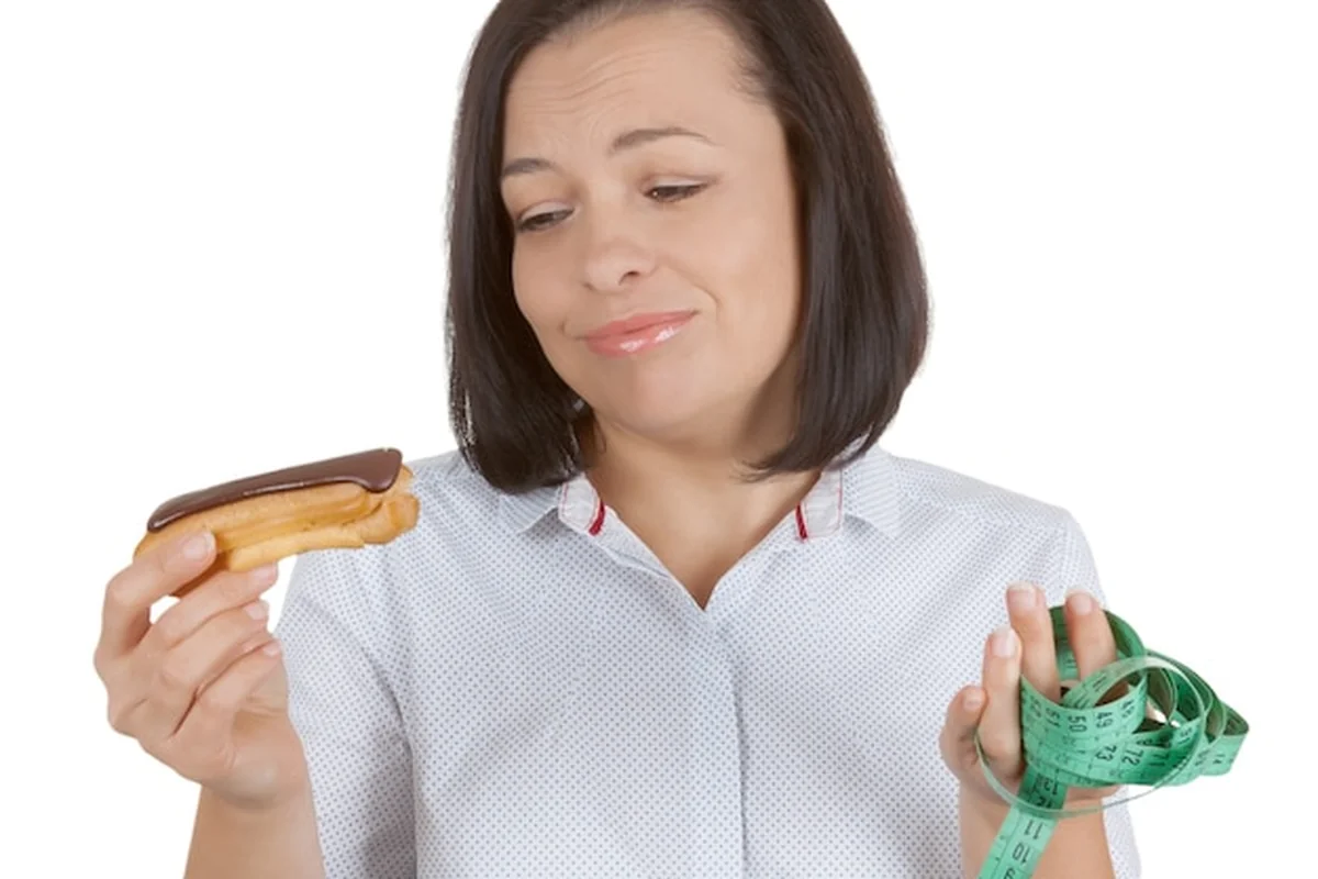 Młoda kobieta wybierająca między czekoladowym eklerem a zdrowiem na białym tle