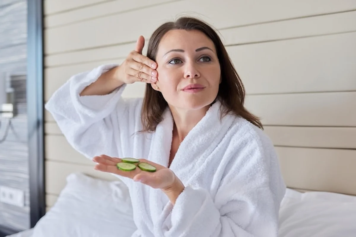 Dojrzała kobieta w szlafroku pielęgnuje skórę twarzy z ogórkami i masażem w łóżku